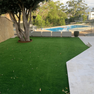 A backyard with artificial grass, a tree, and a swimming pool in the background.